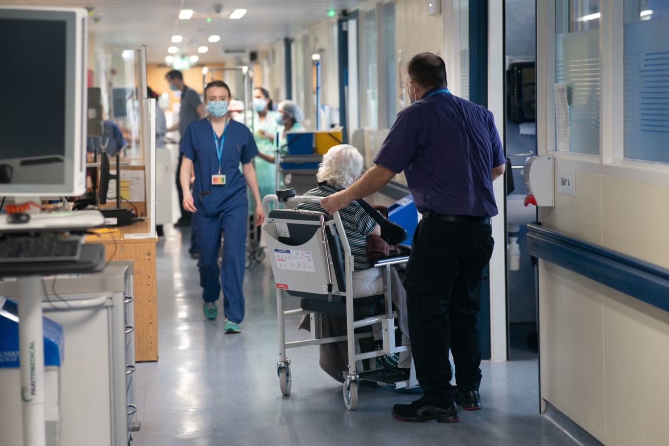 A nurse in scrubs walking down a hospital ward corridor while a porter moves a patient in a wheelchair