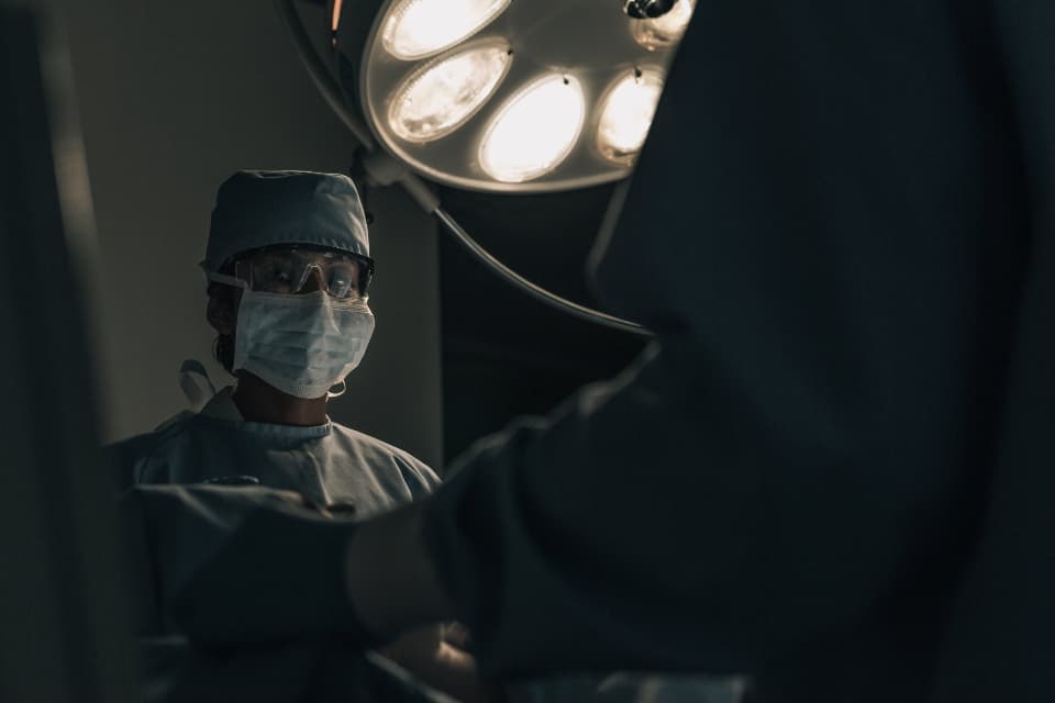 A clinician in scrubs and a surgical mask under the lights of an operating theatre